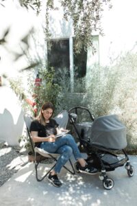 A woman sits reading next to a baby stroller in a sunny outdoor setting.