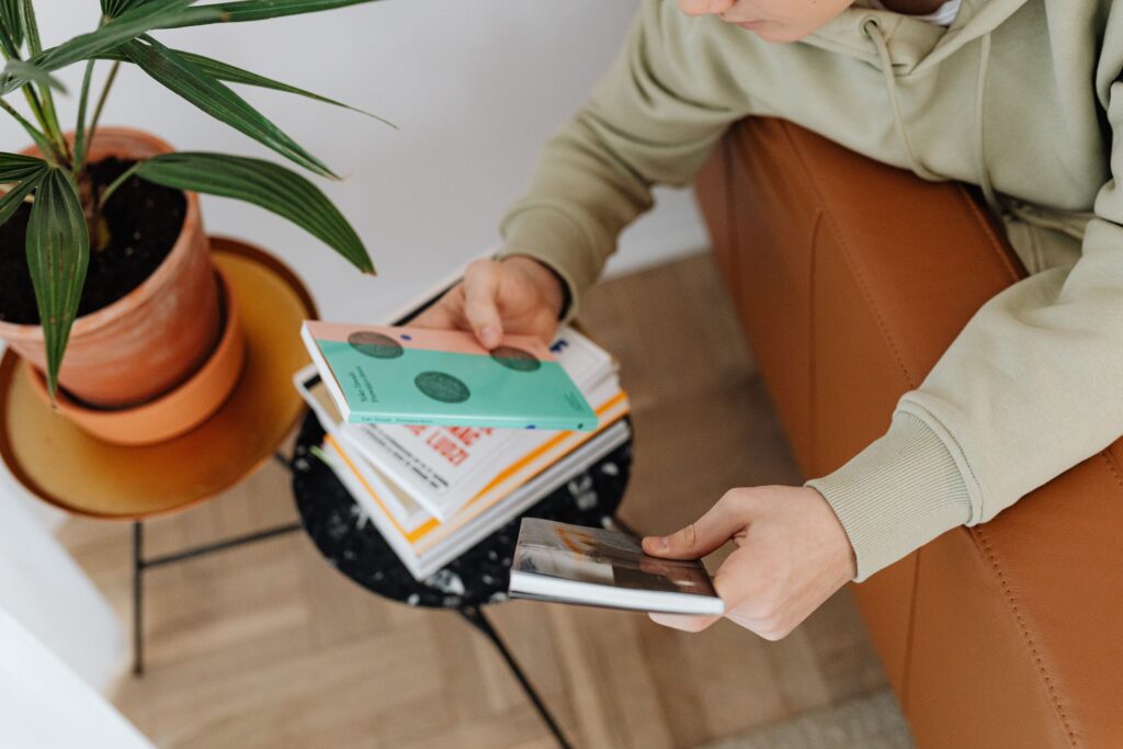 Man casually selecting books from a stack while sitting on a leather sofa indoors.