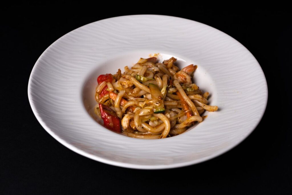 Close-up of stir-fried udon noodles with vegetables on a white plate against a dark background. Udon: macarrão grosso.